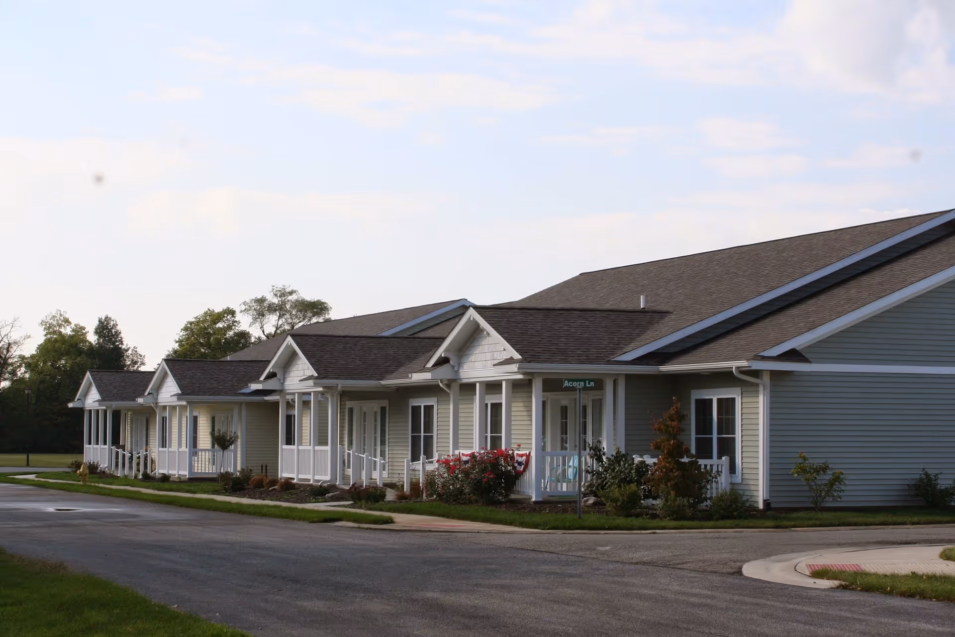 Exterior view of a single-story residential building with multiple units, each having a small porch with white railings. The building is light-colored with a dark roof, surrounded by greenery and a paved road in front.