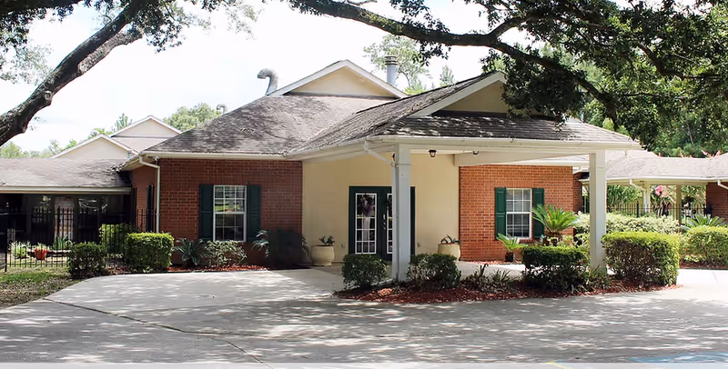 Front exterior of a single-story brick building with a covered entrance, driveway, and landscaped shrubs under large trees.