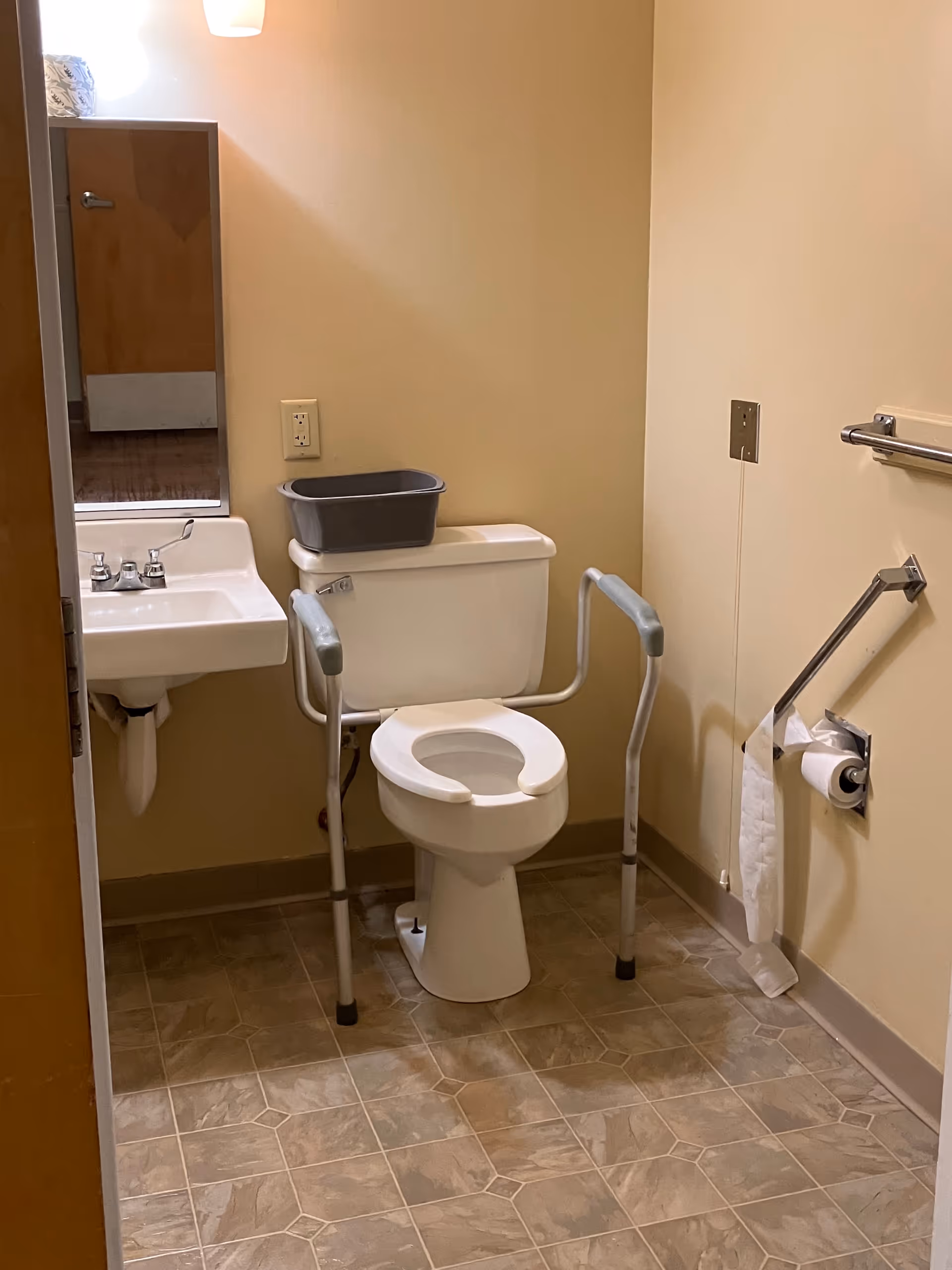 Accessible bathroom with toilet, sink, mirror, grab bars, and a bucket on the toilet.