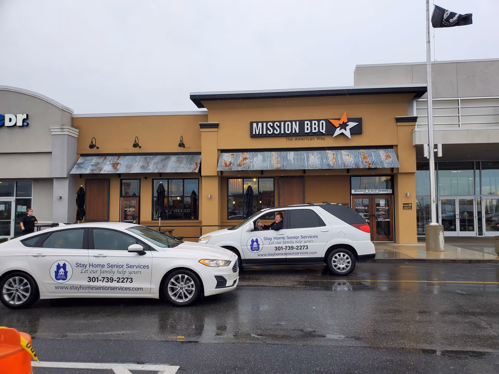 Two cars with Stay Home Senior Services logos parked in front of a storefront strip mall.