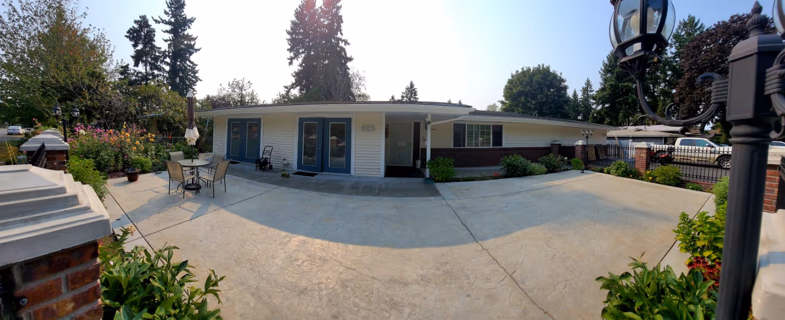 Wide exterior view of a single-story adult family home with a concrete patio area. The house has white siding with blue doors and windows, surrounded by greenery including trees, bushes, and flowers. There is a small outdoor seating area with a table and chairs on the left side. A black metal fence and a parked white vehicle are visible on the right side.