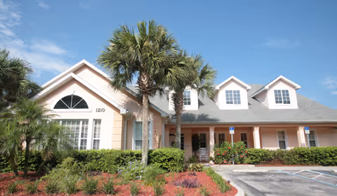 Exterior front view of a single-story residential building with a light peach facade, multiple windows, and a gray shingled roof. There are palm trees and well-maintained shrubs in front of the building, along with a parking area that includes handicap parking spaces. The sky is clear and blue.