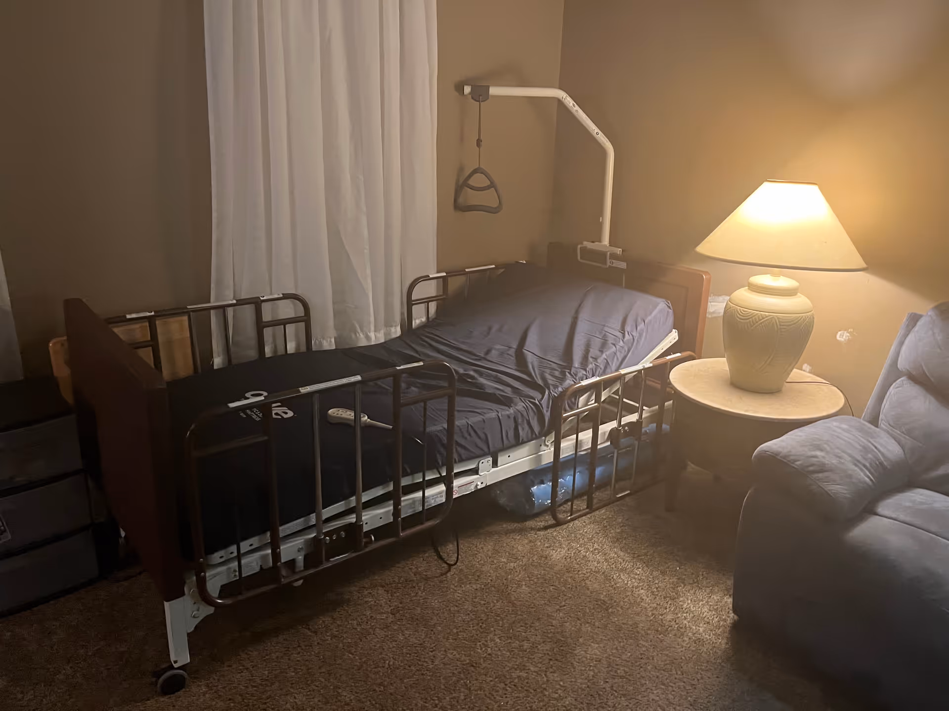 Hospital-style adjustable bed in a small bedroom with a nightstand and lamp.