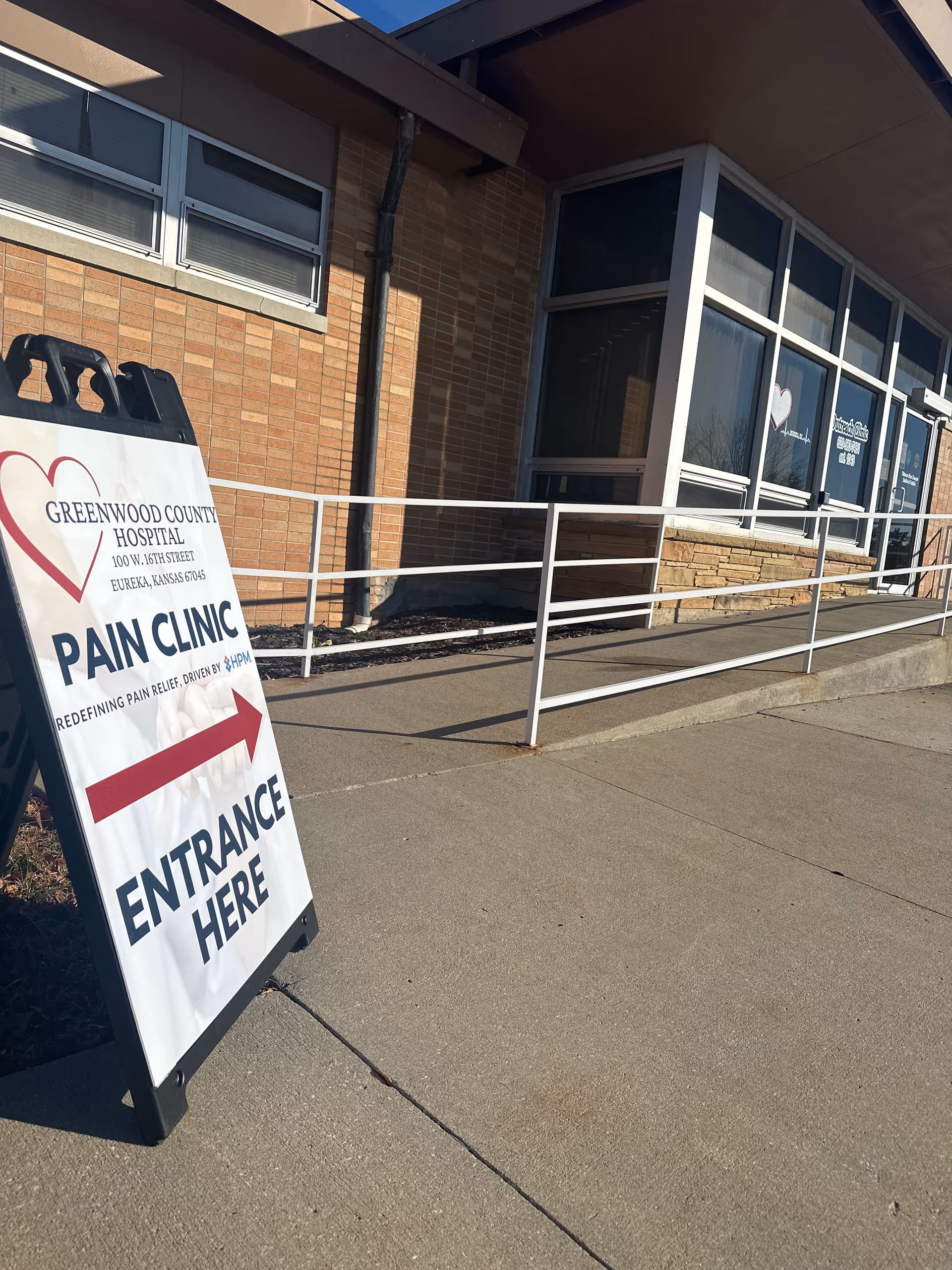Exterior view of Greenwood County Hospital entrance with a large pain clinic sign on the sidewalk.