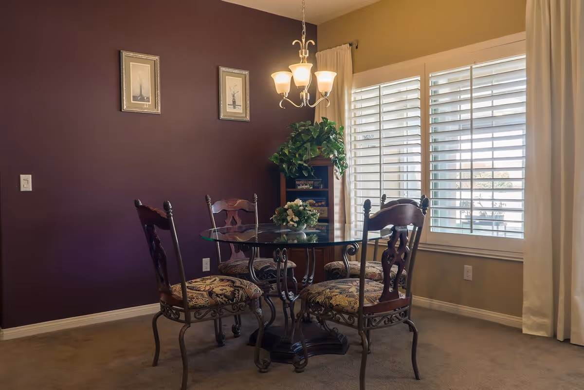A dining area with a round glass table surrounded by four ornate chairs with patterned cushions. The room has a purple accent wall with two framed pictures, a window with white shutters and curtains, a small bookshelf with a green plant on top, and a chandelier with five lights hanging above the table.