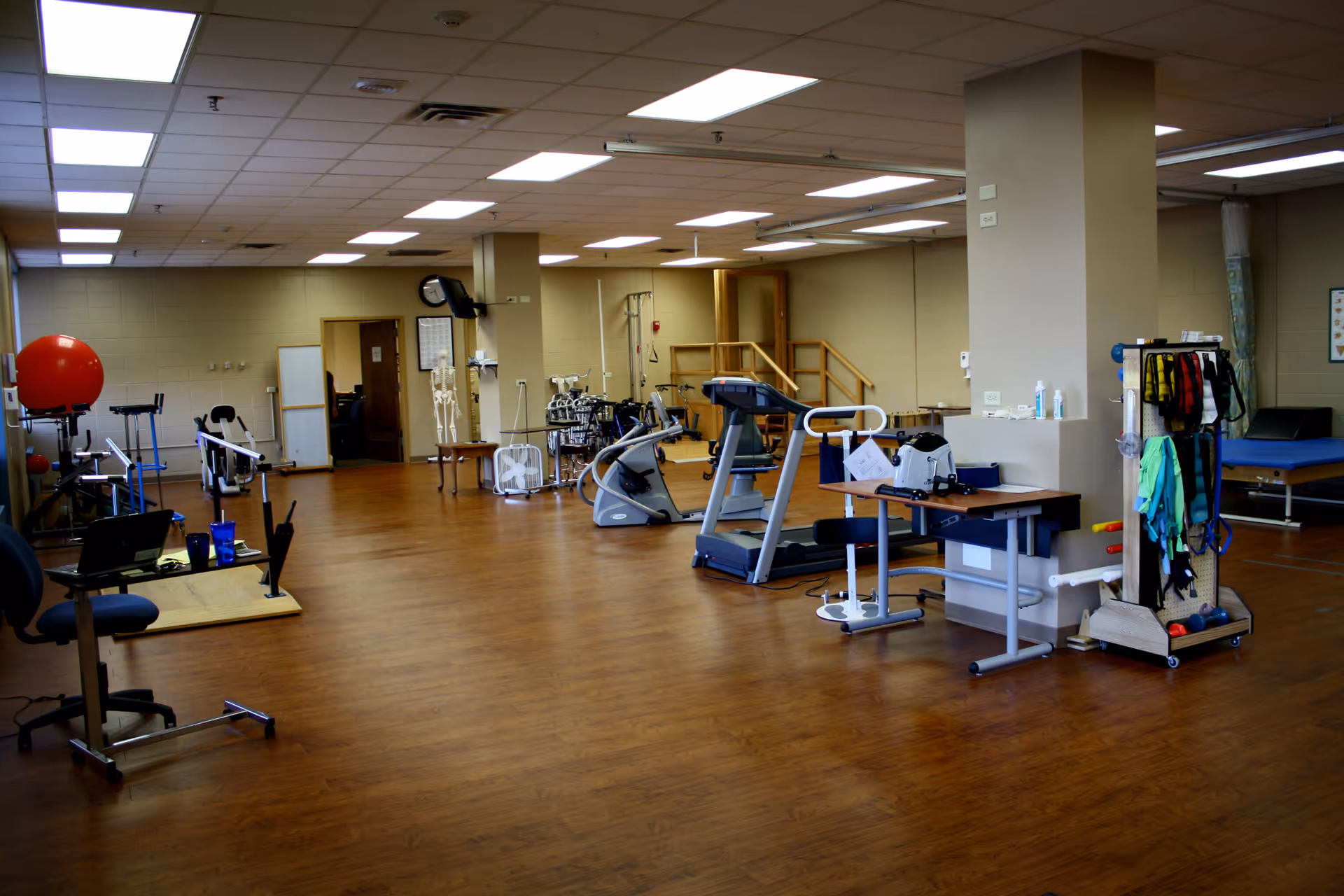 A spacious rehabilitation or physical therapy room with wooden flooring, exercise equipment including a treadmill, stationary bike, parallel bars, therapy tables, and various therapy tools and supplies. The room is well-lit with ceiling lights and has beige walls.