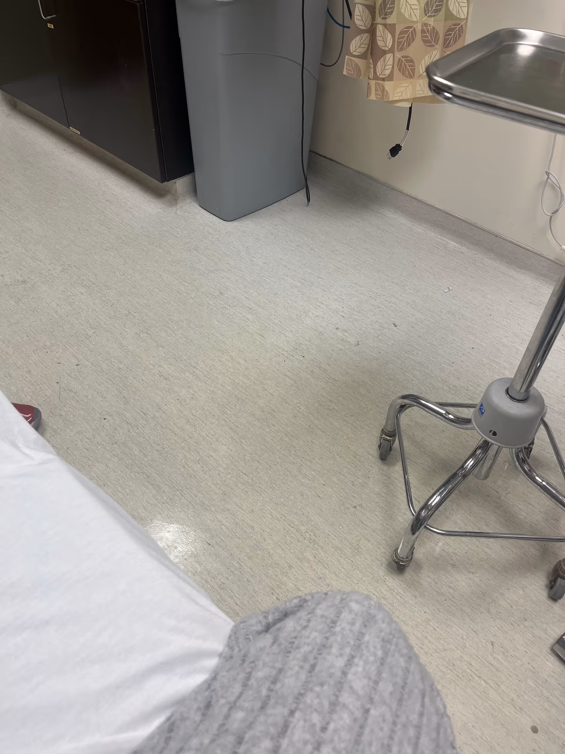 A clinical room with a bed, curtain, gray cabinet, and wheeled medical stand on linoleum floor.