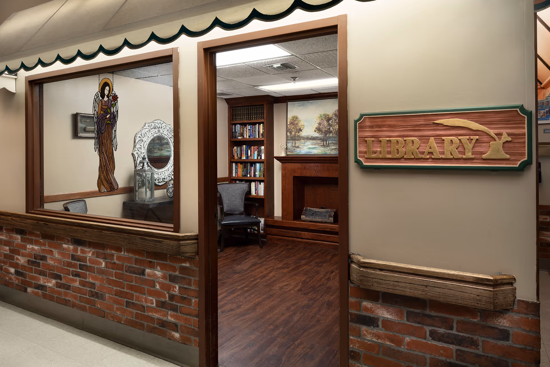 View of a library room in a senior living facility with a wooden floor, a bookshelf filled with books, a chair, a fireplace, and a painting of trees above the fireplace. The entrance has a wooden sign that reads 'LIBRARY' with a quill design. There is a window with a stained glass angel decoration and a decorative mirror inside the room.