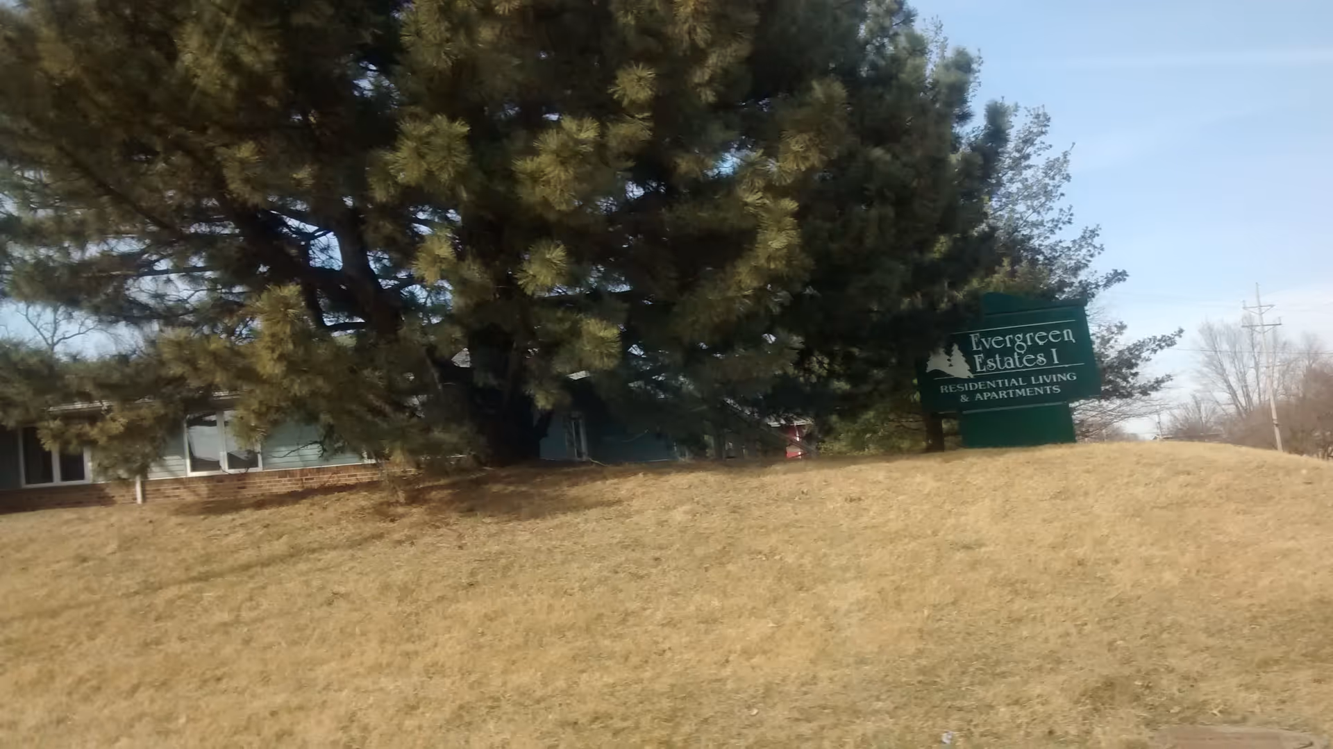 Exterior view of Evergreen Estates I sign on a grassy slope with a building and trees.