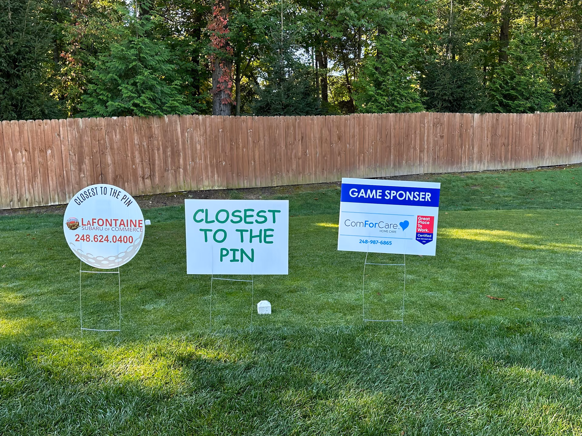 Several signs on a grassy lawn in front of a wooden fence advertising sponsors.