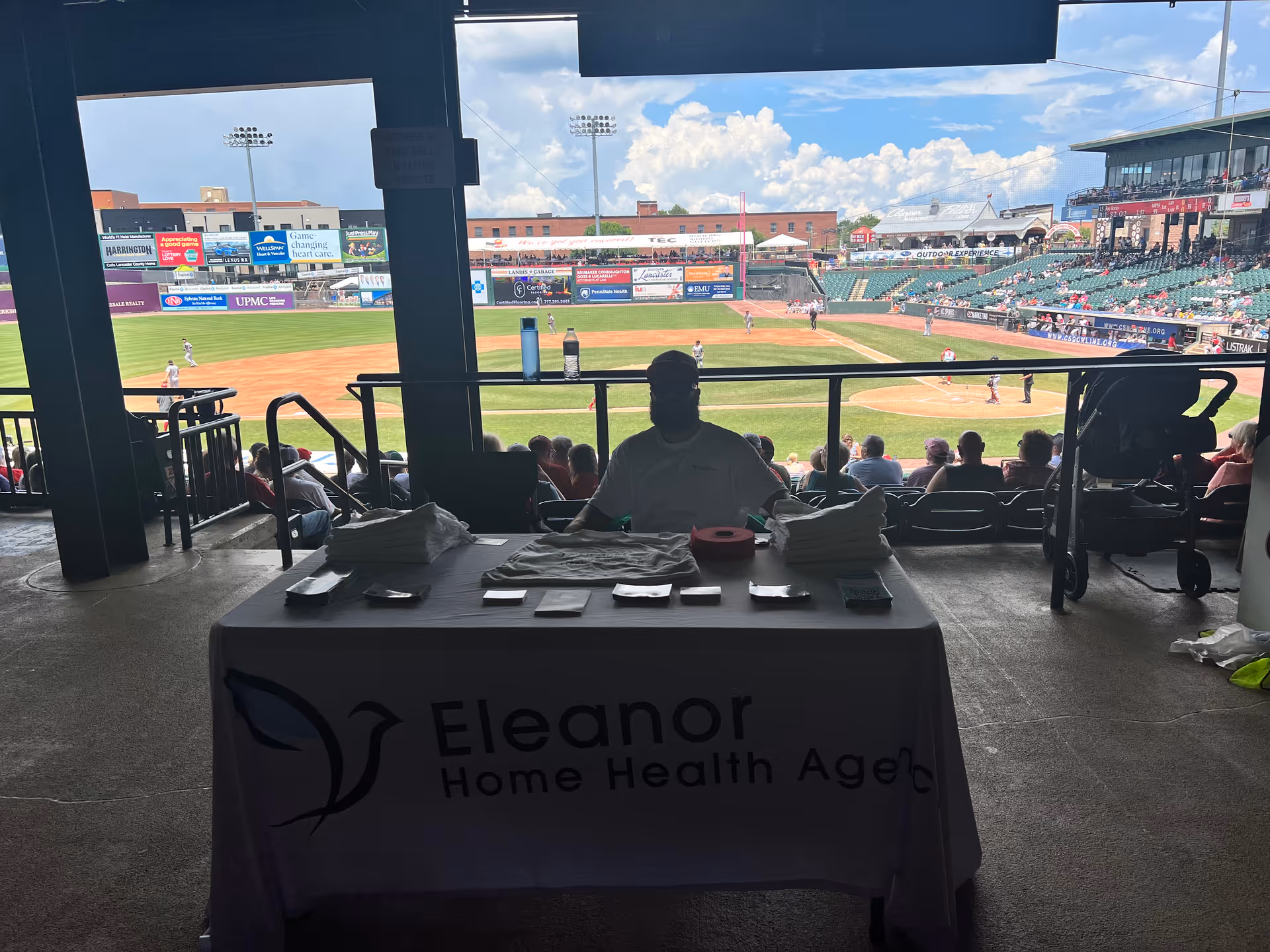 Outdoor stadium concourse with a table displaying Eleanor Home Health Agency banner and attendees.