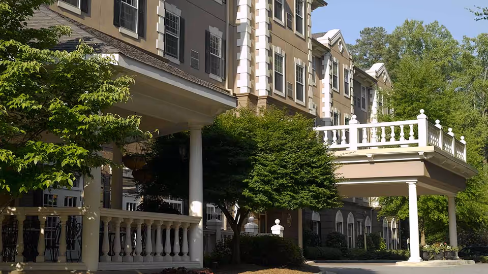 Exterior view of a multi-story residential building with beige walls, white trim, and multiple windows. The building features a covered entrance supported by white columns and a white railing above. There are green trees and shrubs around the entrance area under a clear blue sky.