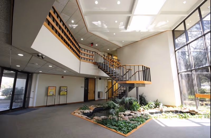 Bright two-story lobby with a curved staircase, plants, and water feature inside a senior living facility.