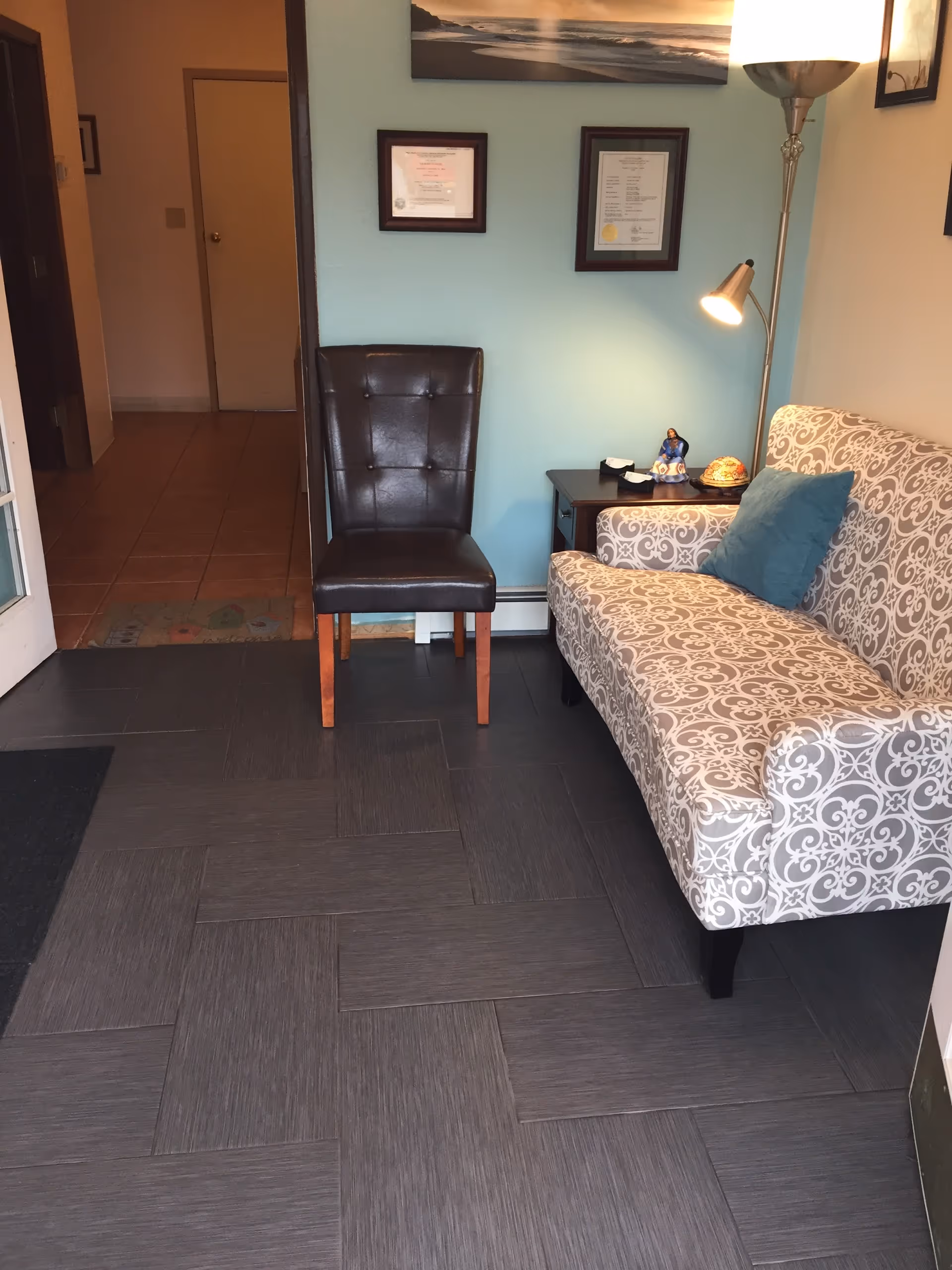 Interior waiting area with a patterned sofa, black chair, side table, and certificates on teal wall.