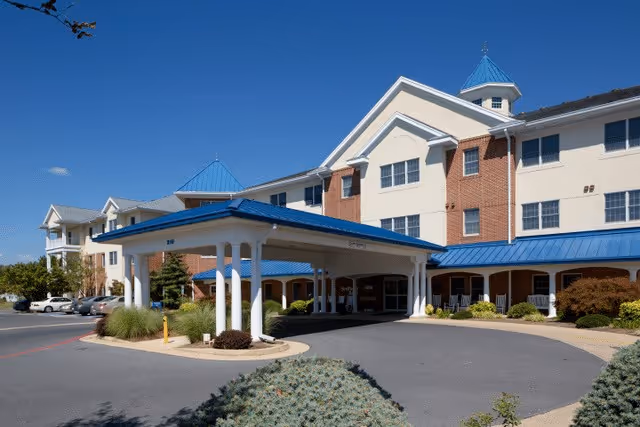 Front entrance of a multi-story retirement community building with a covered porte-cochère and blue roofs under a clear sky.