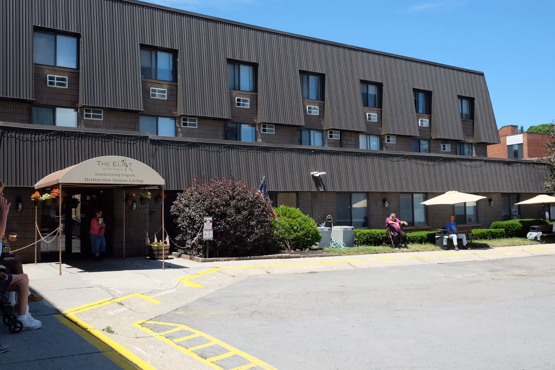 Exterior view of The Eliot Erie Station assisted living facility with a brown multi-story building, an entrance canopy, some bushes, and a few people sitting outside under umbrellas on a sunny day.
