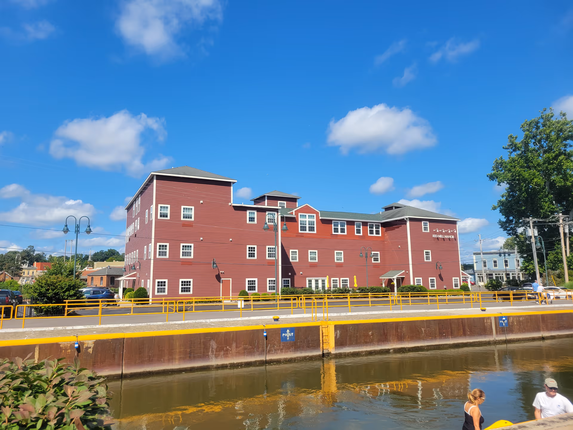 Red three-story senior living building along a canal with a yellow railing and blue sky.