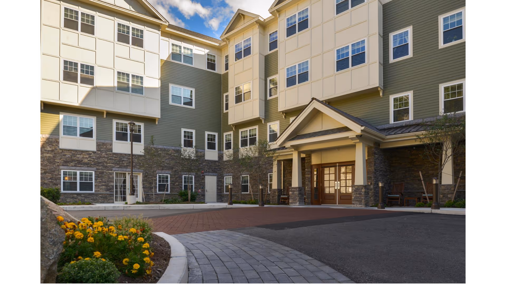 Exterior view of a senior living facility building with multiple windows, a covered entrance with double doors, stone and siding facade, a paved driveway, and landscaped flower beds with yellow flowers.