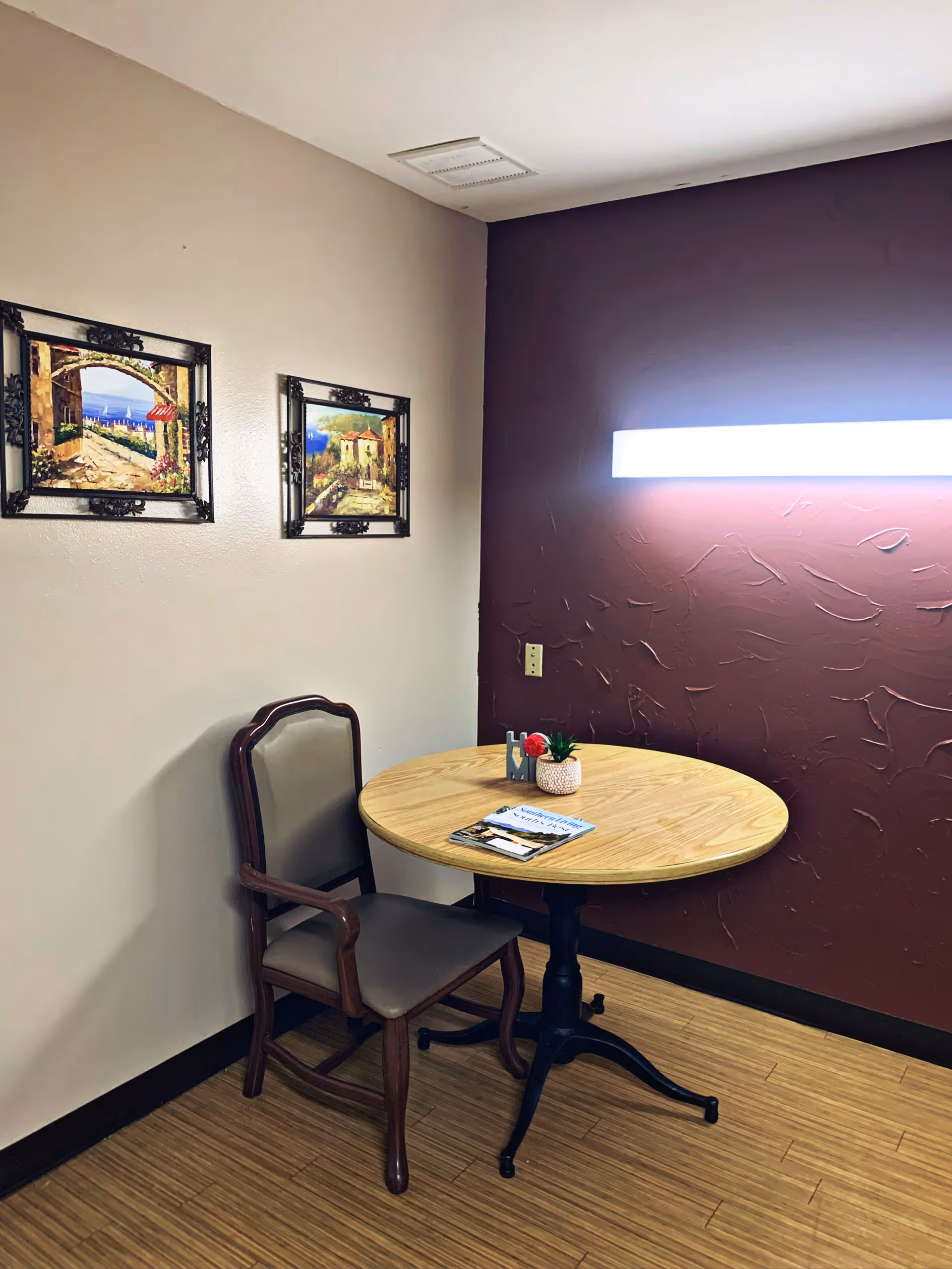 A small corner of a room with a round wooden table and a single wooden chair with padded seat and backrest. On the table, there is a small potted plant, a decorative item spelling 'HOME', and a magazine. The walls are painted two different colors: one beige and one dark maroon with textured patterns. Two framed paintings depicting outdoor scenes hang on the beige wall. A bright rectangular light fixture is mounted on the maroon wall above the table.