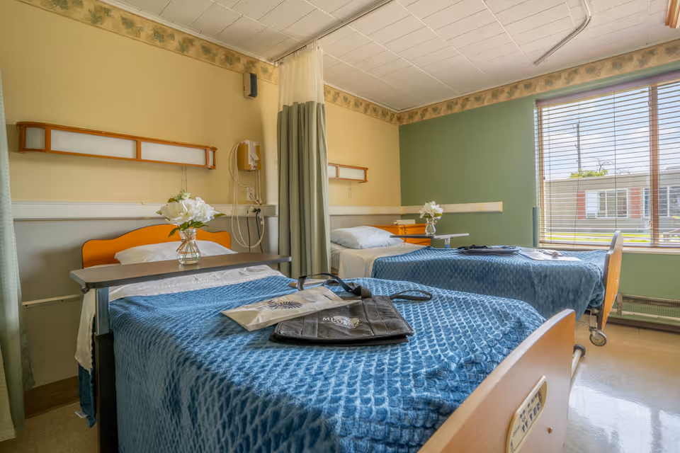 A nursing facility room with two hospital beds covered in blue blankets. Each bed has a small table with a vase of white flowers. There is a large window with blinds letting in natural light, and a curtain divider between the beds. The walls are painted in light yellow and green with a floral border near the ceiling.