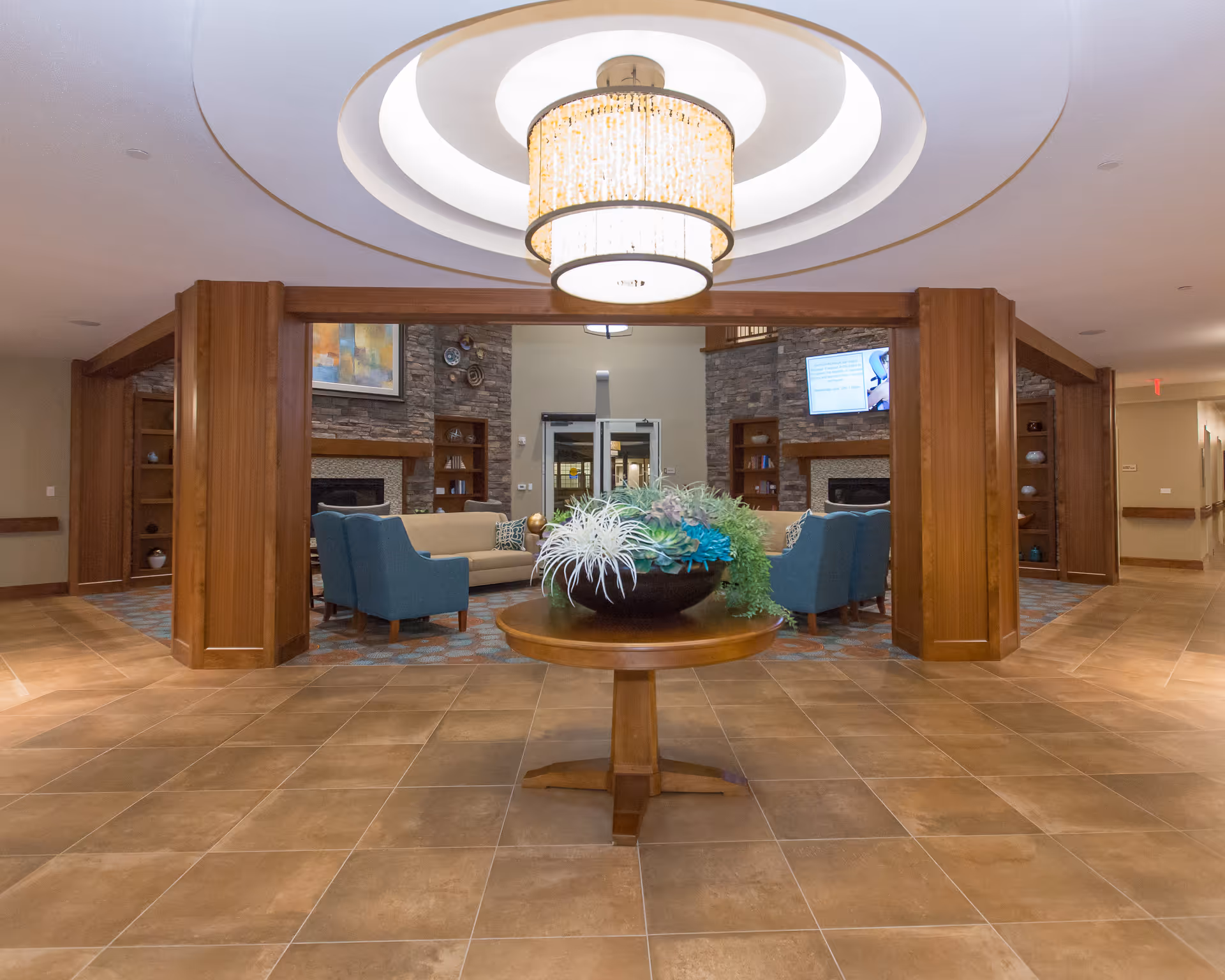 Spacious senior living facility common area with a round wooden table holding a large decorative plant in the center. The room features tiled flooring, wooden pillars, and a modern chandelier hanging from a recessed ceiling. In the background, there are comfortable blue and beige chairs arranged around two stone fireplaces with built-in wooden shelves and a wall-mounted TV.