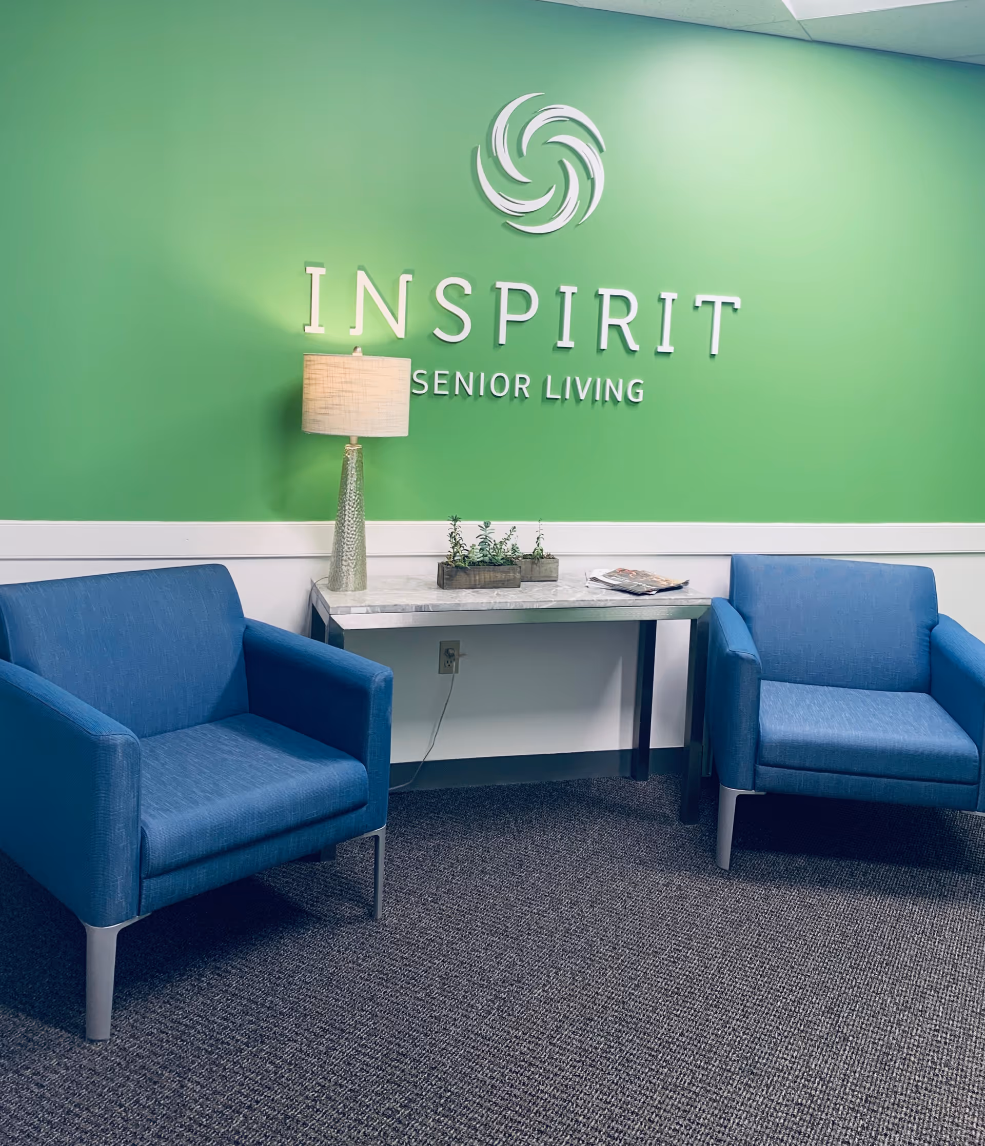Green wall with Inspirit Senior Living logo, blue chairs, lamp, plants, and table.