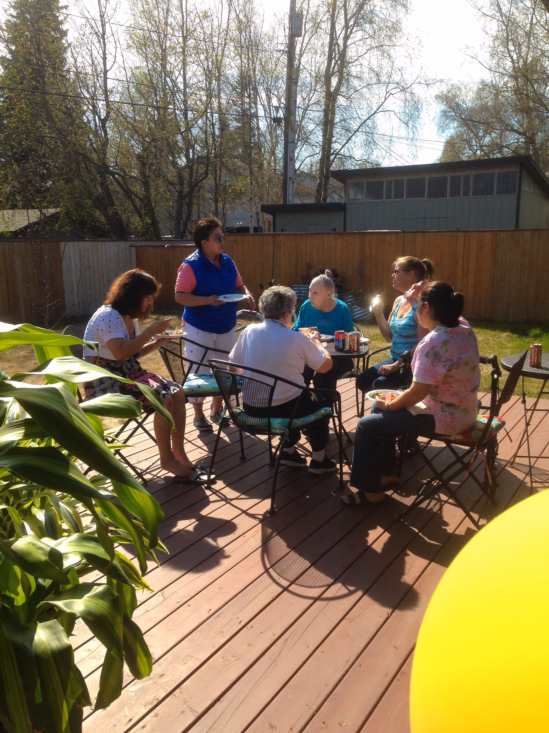 A group of six people sitting and standing around a table on a wooden deck outdoors, eating and drinking on a sunny day. There are trees and a wooden fence in the background.