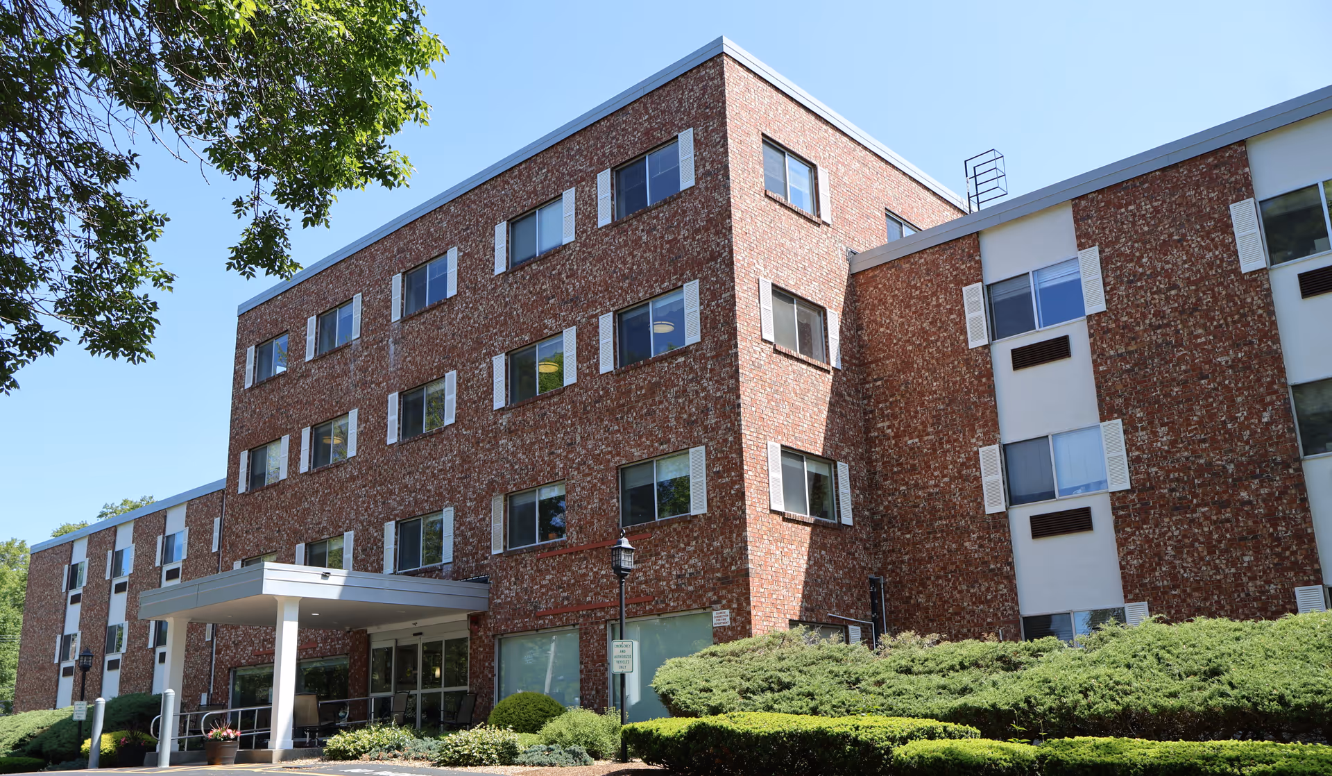 Exterior view of a multi-story brick building with numerous windows, surrounded by green bushes and trees under a clear blue sky. The building has a covered entrance with a ramp and a lamppost nearby.