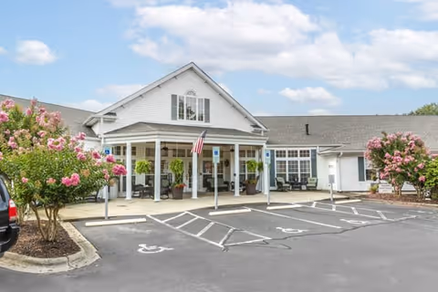 Front exterior view of Brookdale Reidsville facility showing a white building with a covered entrance, an American flag, several potted plants, and pink flowering bushes on either side. There are multiple handicap parking spaces in front of the entrance under a partly cloudy sky.