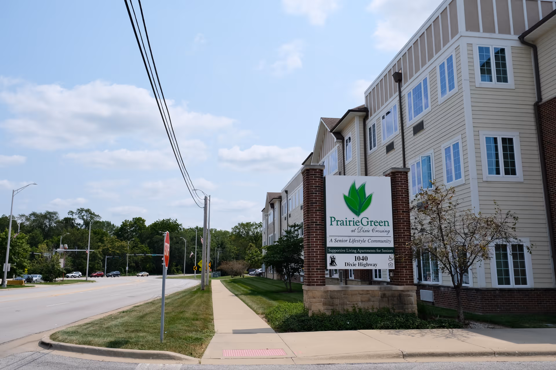 Front exterior of the Prairie Green at Dixie Crossing senior living building with a large sign by the sidewalk next to a road.