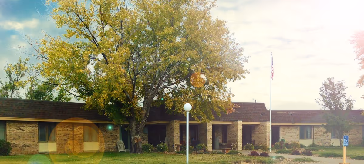 Front view of a single-story brick retirement village building with a large tree, flagpole, and lawn.