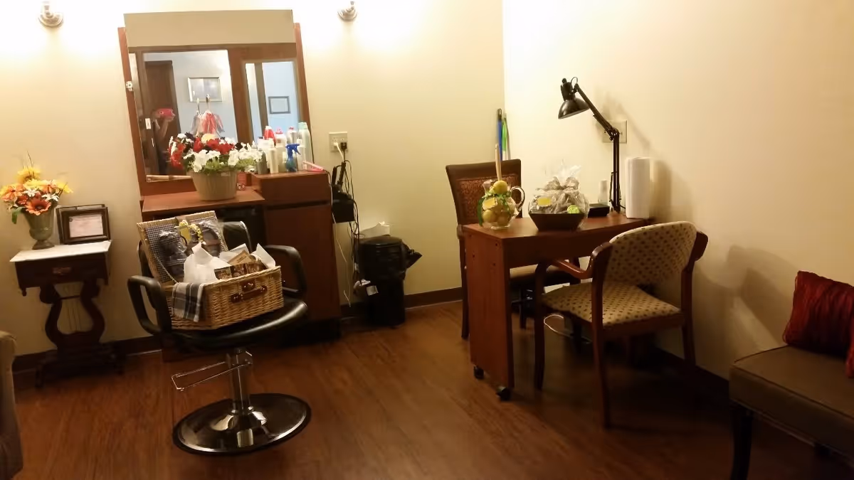 A small room with wooden flooring featuring a black swivel chair with a basket on it, a wooden vanity with a mirror and various bottles on top, a small side table with a flower arrangement, and a wooden desk with two chairs. The desk has decorative items and a black desk lamp. The walls are light-colored and there is a cushioned bench with a red pillow on the right side.