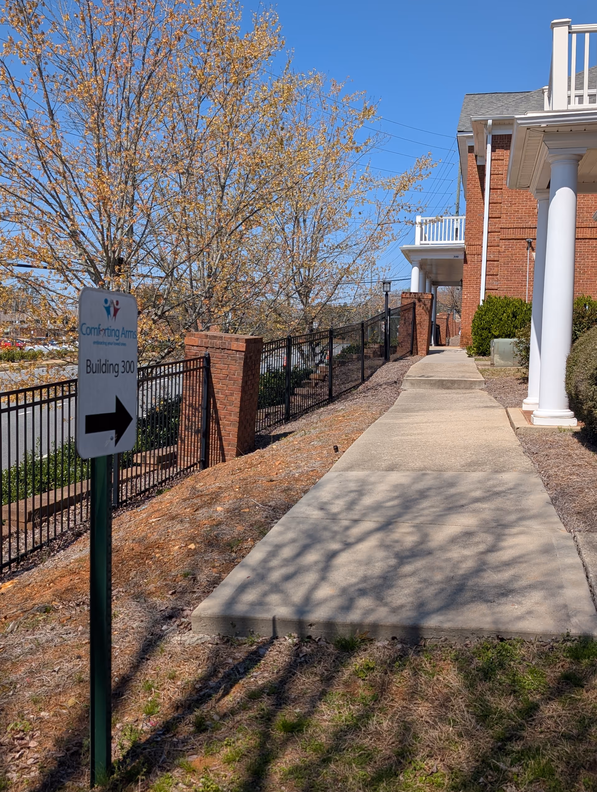 Sidewalk leads to Comforting Arms entrance beside brick building and yellow-leaved trees.