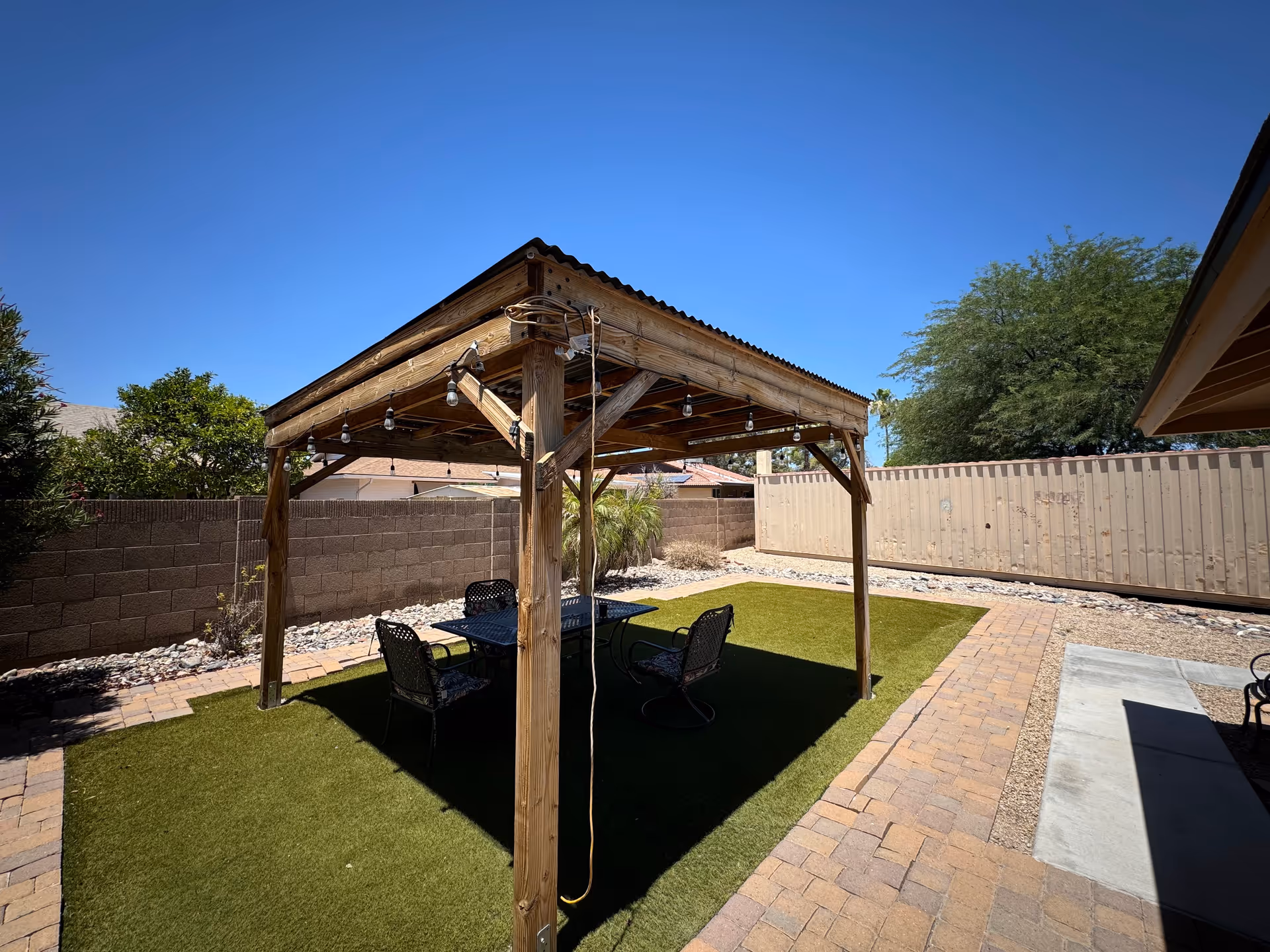 Outdoor patio area with a wooden pergola covering a table and four chairs on artificial grass, surrounded by a brick pathway and a fenced yard with trees and shrubs under a clear blue sky.
