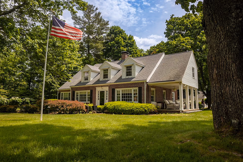 A large brick and white siding house with a sloped roof and dormer windows, surrounded by green trees and bushes. An American flag on a flagpole stands on the lawn in front of the house. The house has a covered porch with outdoor seating and a well-maintained grassy yard.
