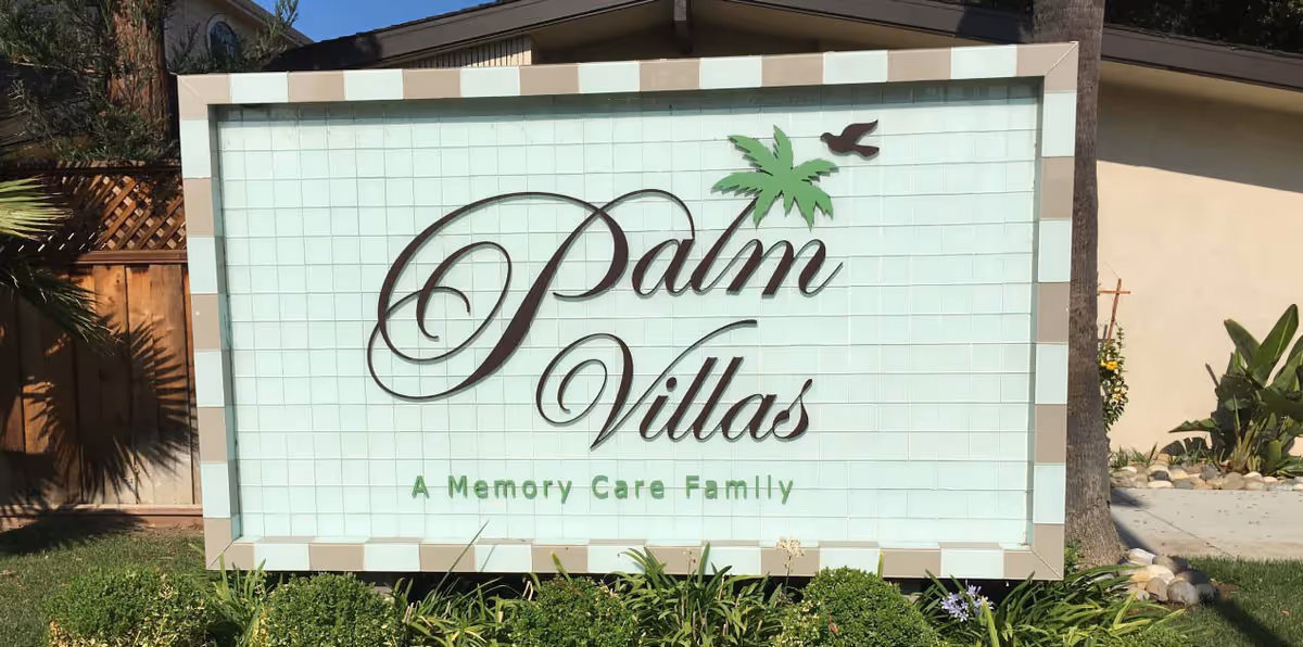 A large outdoor sign reading "Palm Villas" with a palm tree logo and the subtitle "A Memory Care Family" surrounded by landscaping.
