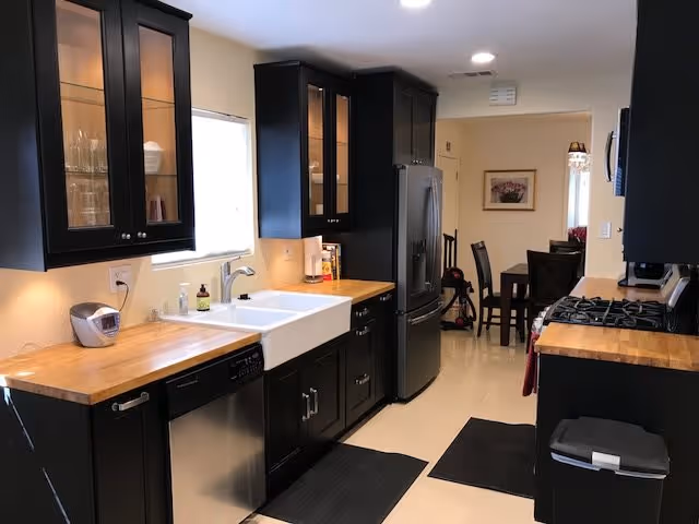 Modern kitchen with black cabinets, wooden countertops, a white farmhouse sink, stainless steel dishwasher and refrigerator, and a gas stove. The kitchen opens into a dining area with a dark wooden table and chairs. The space is well-lit with recessed ceiling lights and a window above the sink.