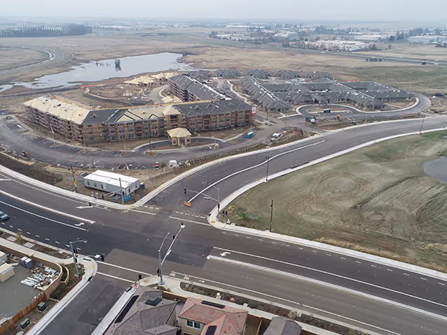 Aerial view of a large assisted living and memory care facility under construction, with multiple connected buildings, surrounding roads, and open land in the background.
