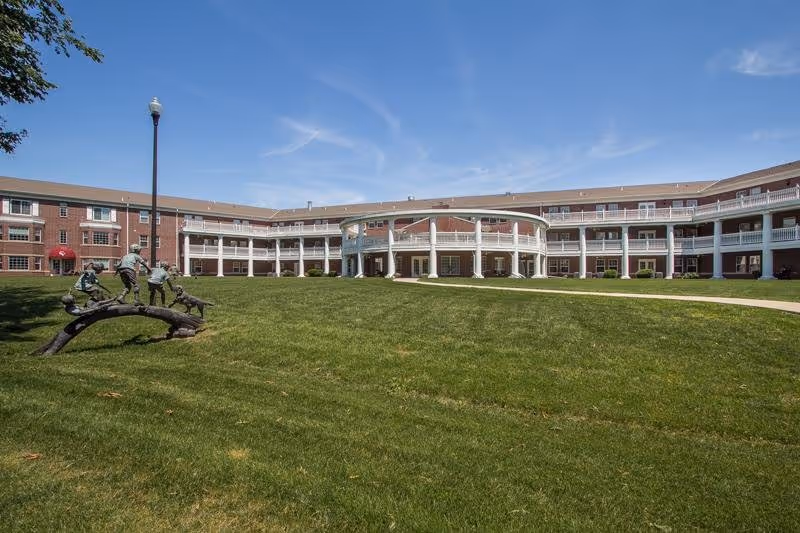 Sunny exterior view of Carnegie Village Senior Living Community with a curved central entrance and lawn.