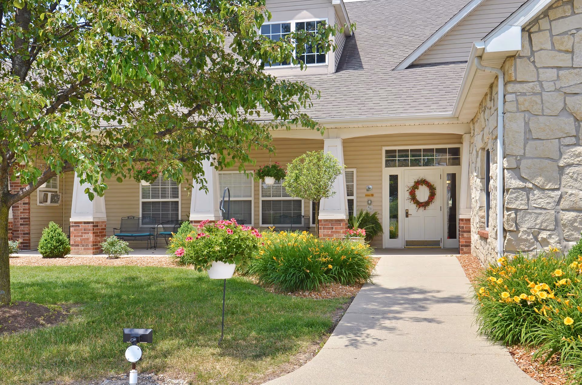 Entrance of a senior living facility with a concrete walkway leading to a white door decorated with a wreath. The building exterior features beige siding, stone walls, and brick accents. There are green shrubs, flowering plants, and a tree in the landscaped front yard.