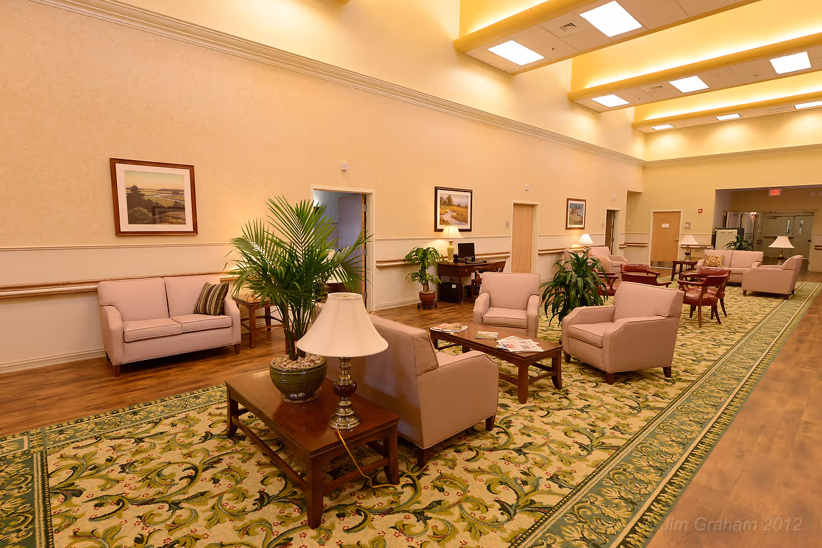 A spacious and well-lit common area with beige walls and a patterned green carpet. The room is furnished with multiple beige armchairs and sofas arranged around wooden coffee tables. There are several potted plants and table lamps placed on side tables. The ceiling has recessed lighting and skylights, and framed landscape paintings hang on the walls.