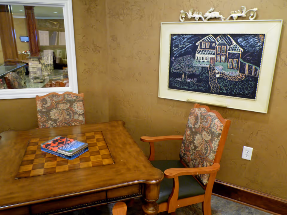 A small room with a wooden table featuring a built-in checkerboard and two upholstered chairs with patterned fabric. On the table are two board games. A framed chalkboard drawing of a house and garden hangs on the textured brown wall. A window shows a view into another room with stone and wood decor.