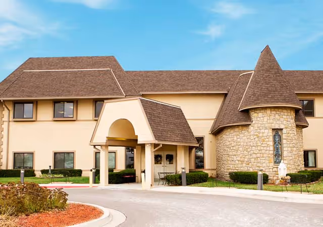 Front exterior of a senior living building showing a covered porte-cochere entrance, a stone turret with a stained-glass window, and a landscaped driveway.