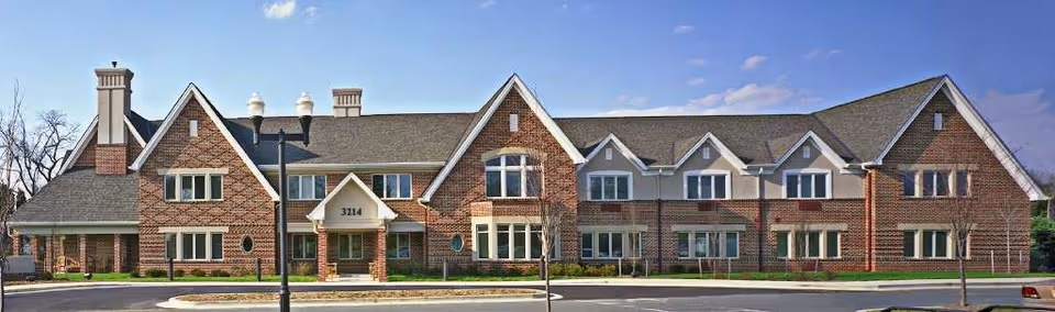 Front exterior view of a two-story brick building with multiple windows, gabled roofs, and chimneys. The building has a central entrance with the number 3214 above it, surrounded by a paved driveway and small landscaped areas with young trees. The sky is clear and blue.