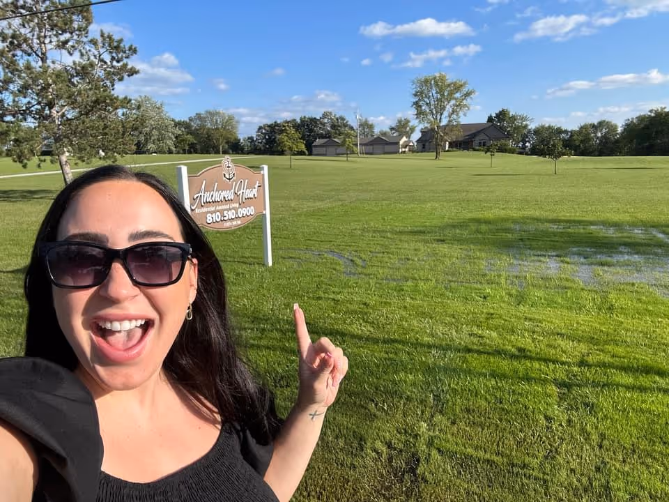 A smiling woman in sunglasses takes a selfie while pointing at a lawn sign that reads "Anchored Heart" in a sunny grassy field with houses in the background.