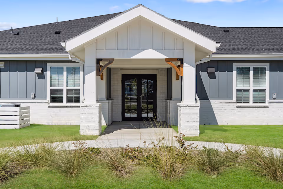 Front exterior view of a single-story building with a covered entrance supported by white columns and wooden brackets. The building has gray siding with white brick on the lower half, black double doors, and windows with white blinds. There is a concrete walkway leading to the entrance, surrounded by green grass and ornamental grasses in the foreground.