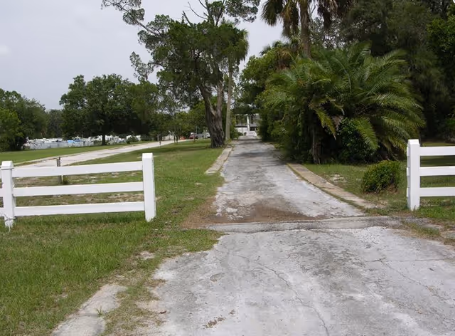 Gravel driveway with white fences leads toward a distant facility surrounded by trees
