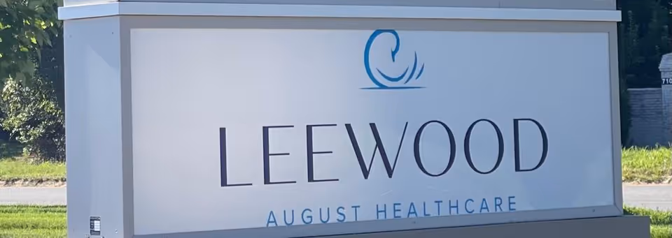 A large white sign with blue and black text that reads 'LEEWOOD AUGUST HEALTHCARE' with a blue circular logo above the text, situated outdoors with grass and trees in the background.