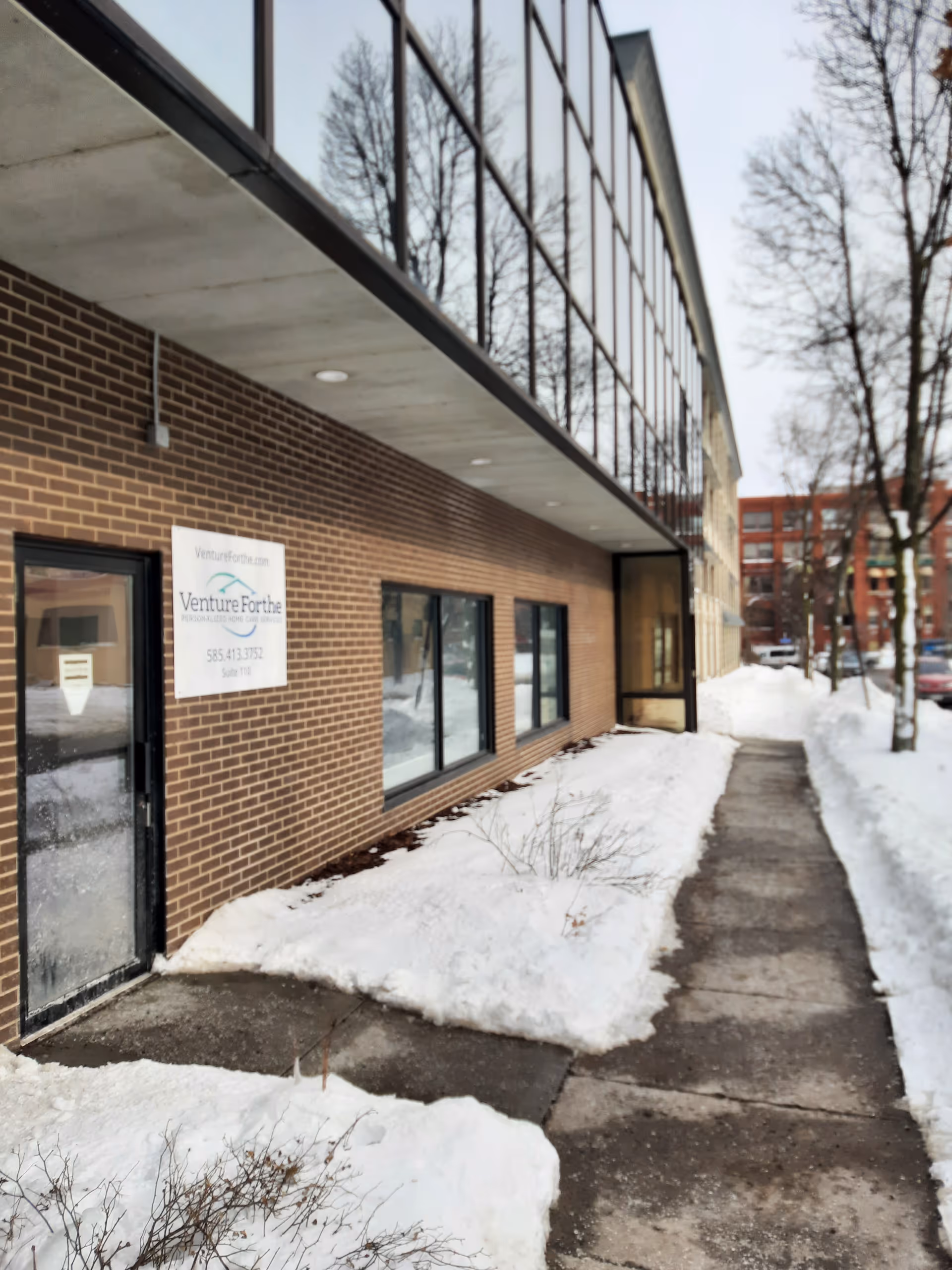 Brick storefront with large windows and a sign for Venture Forthe Rochester, snow along sidewalk.