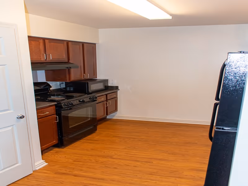 Compact kitchen with wooden cabinets, stove, microwave, and refrigerator in a senior living unit.